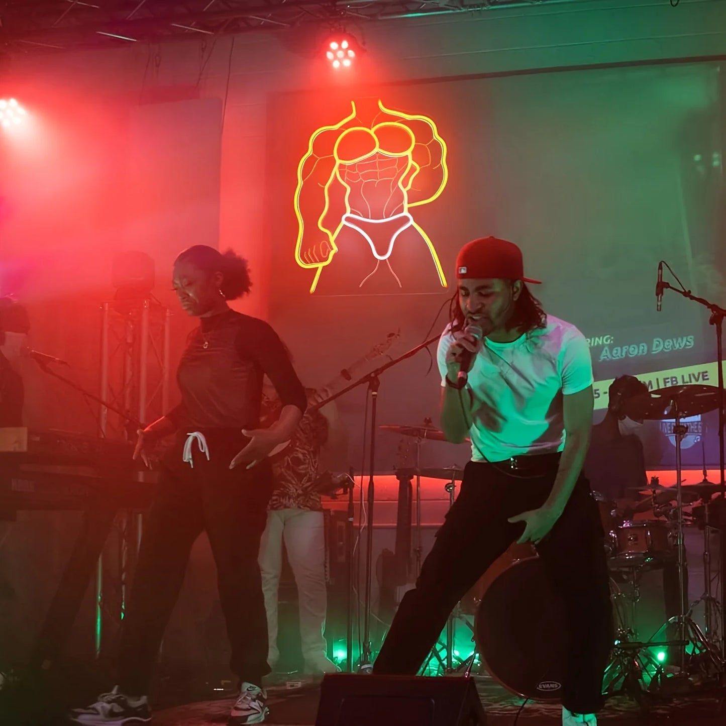 Person on stage with microphone in front of a neon sign and band members in the background.