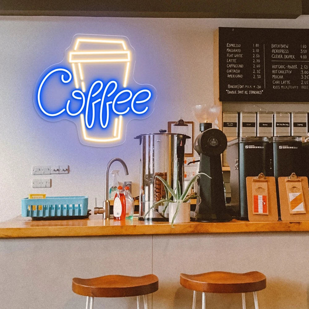 Neon coffee cup sign above a counter with coffee-making equipment and stools in a coffee shop.