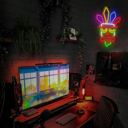 Neon mask light above a computer setup with a colorful screen on a dark wall.
