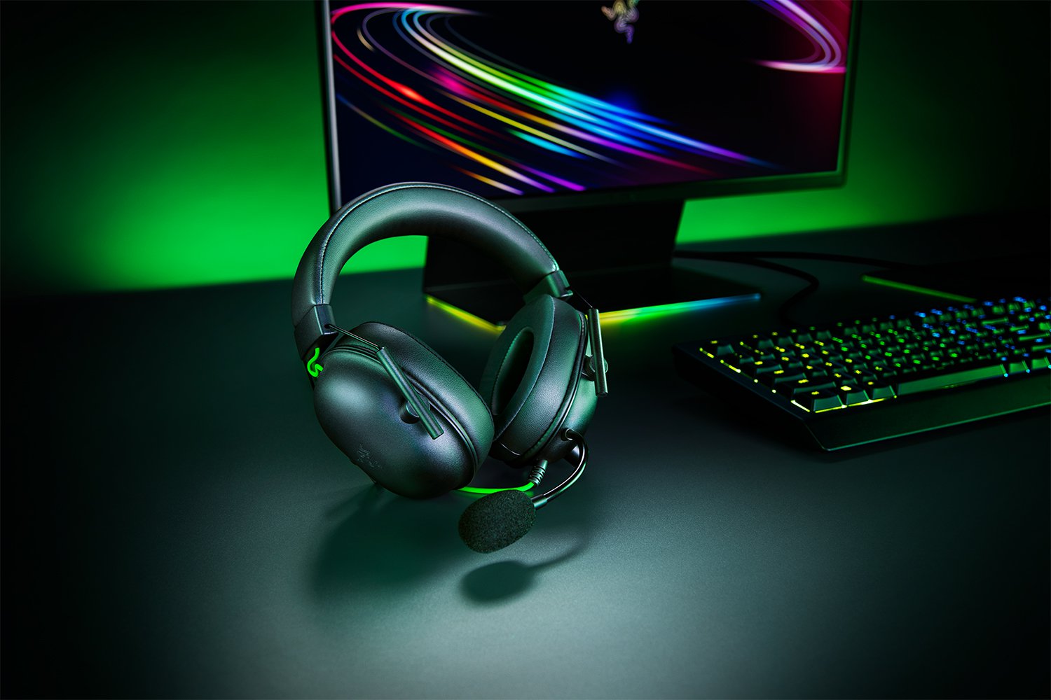 Gaming headset on a desk with a computer monitor and keyboard in a dark room with green lighting.